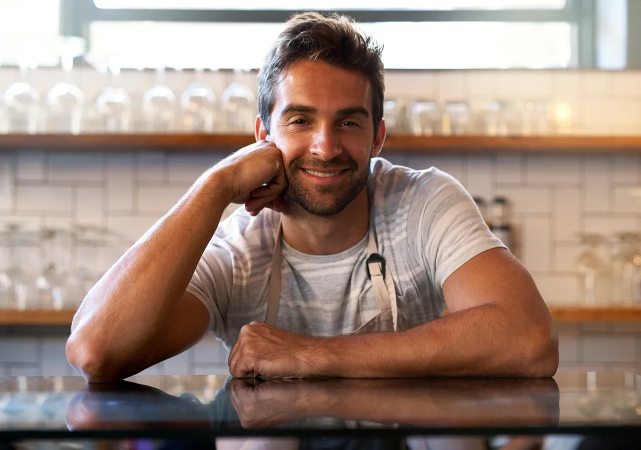 A cafe owner in an apron leaning on the counter of his shop.