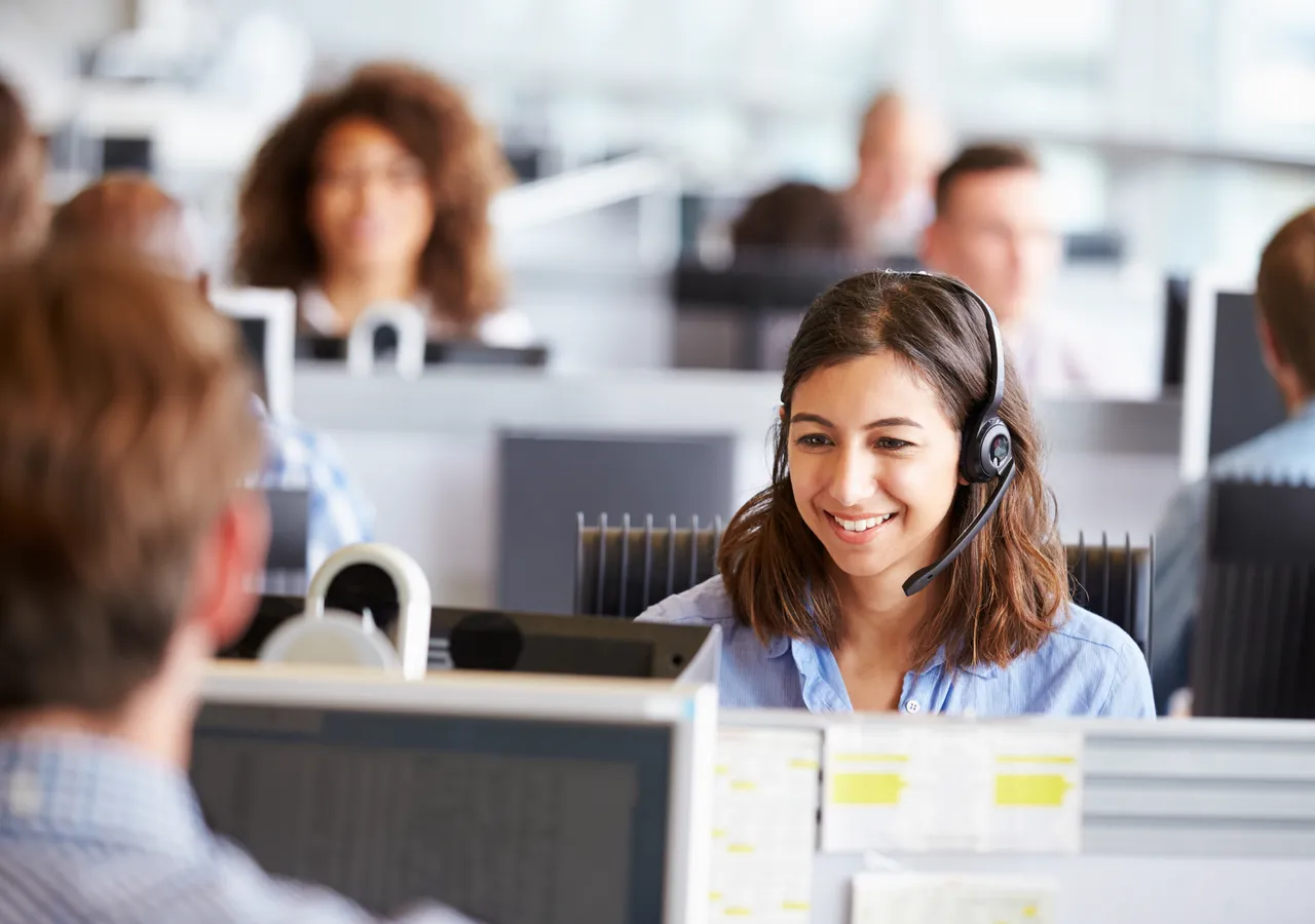 A call centre agent wearing a headset at a workstation, colleagues at desks behind her.