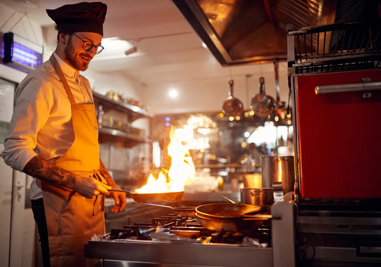 A chef working over a flaming pan on a commercial gas range in a busy professional kitchen, with extraction hoods and equipment around him.