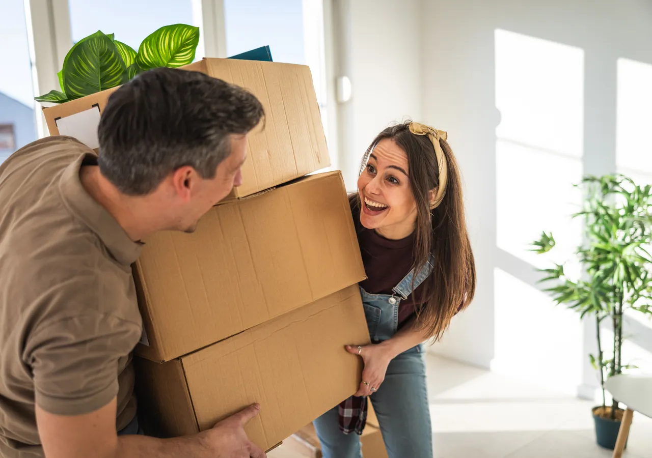 Two people carrying stacked cardboard boxes into an empty room with bare walls and a potted plant beside the door.