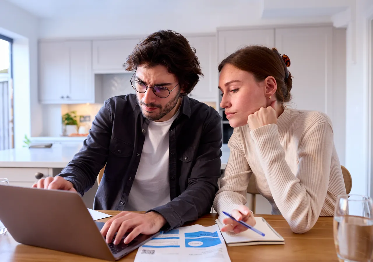 A man and a woman at a kitchen table working through paperwork together with an open laptop and a notepad between them.