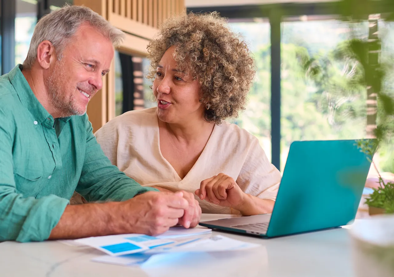 Two people leaning over a laptop at a table, going through printed statements together and pointing at something on the screen.