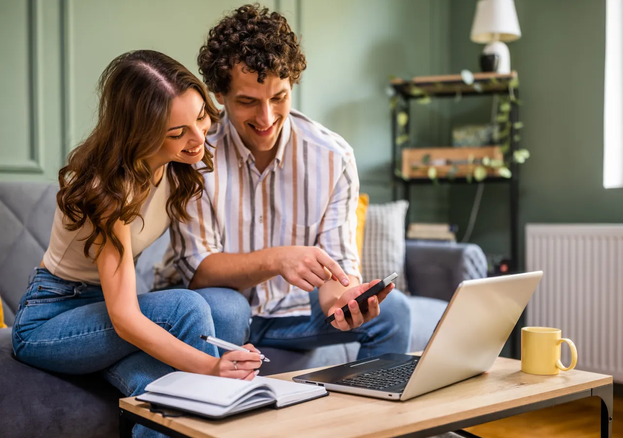 A couple sitting on a sofa with a laptop and a phone, comparing something across both screens.