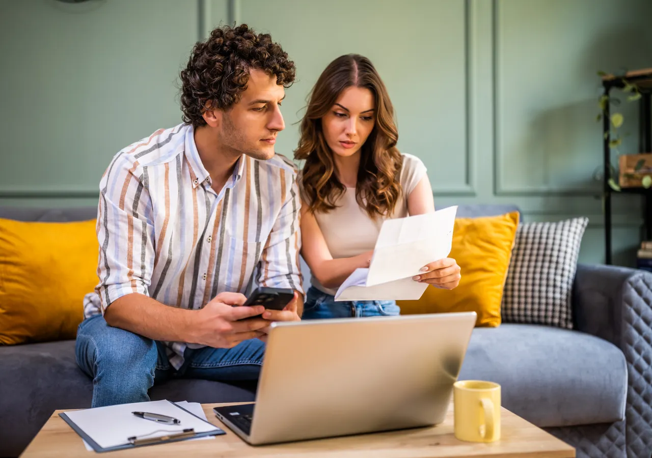 A couple sitting on a sofa with a laptop open in front of them, one holding a printed bill and comparing it against what is on the screen.