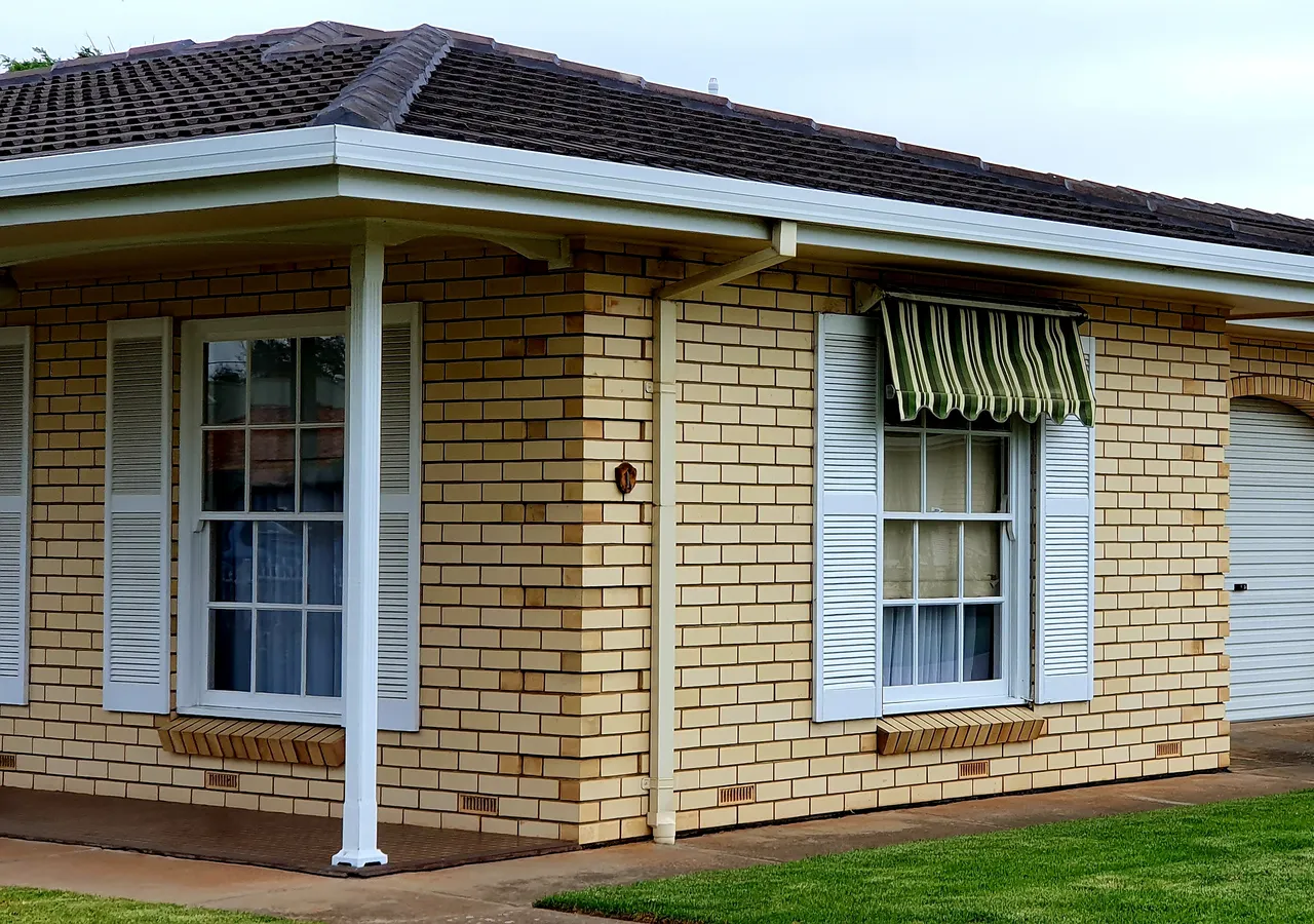 A single-storey Australian cream brick home with a tiled roof, green striped window awnings, white posts and a roller door garage, seen from the front lawn.