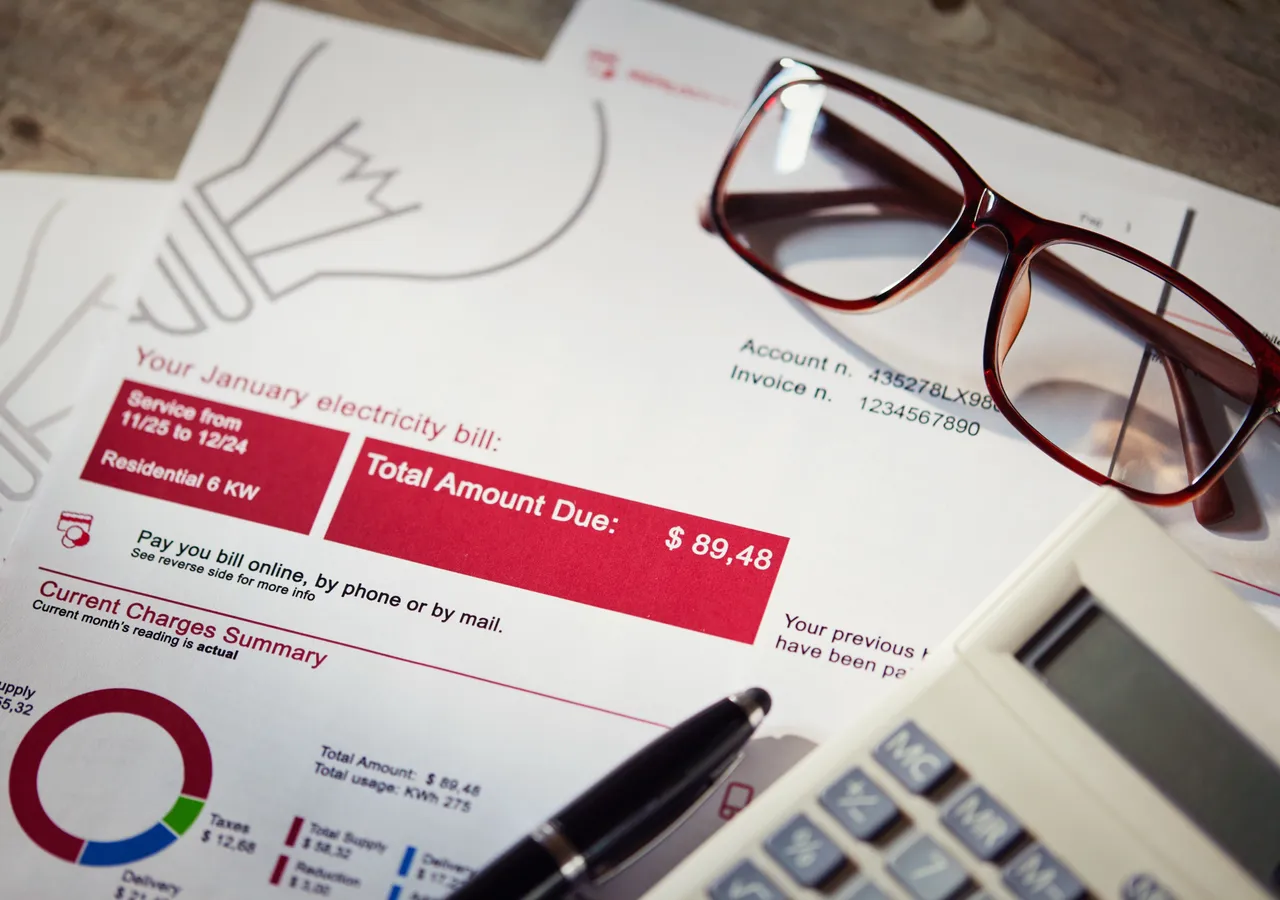 An electricity bill on a desk showing a charges summary and a small pie chart, with a pair of glasses, a pen and a calculator resting on top of it.
