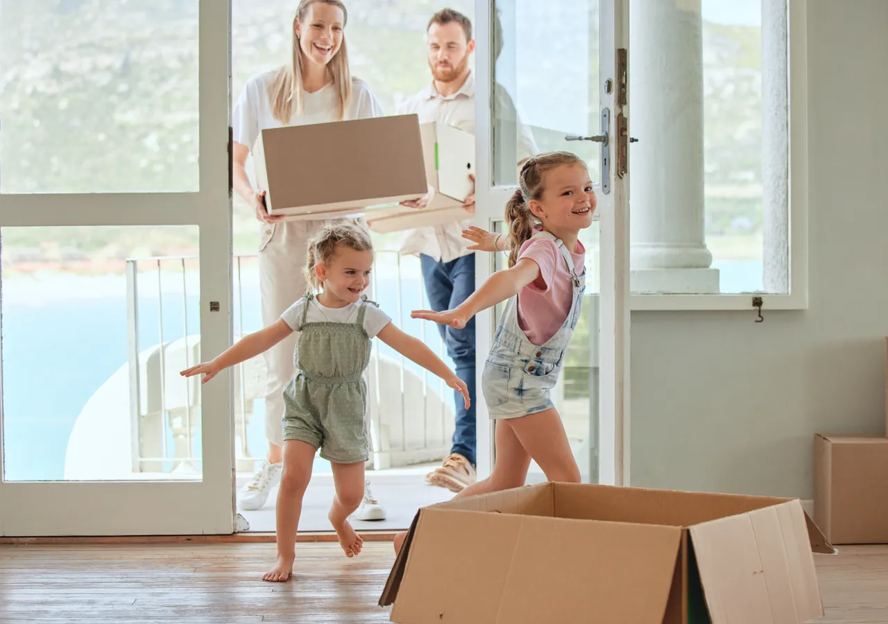 A family carrying cardboard moving boxes through the doorway of a bright, empty room, with two young children running ahead of the adults.