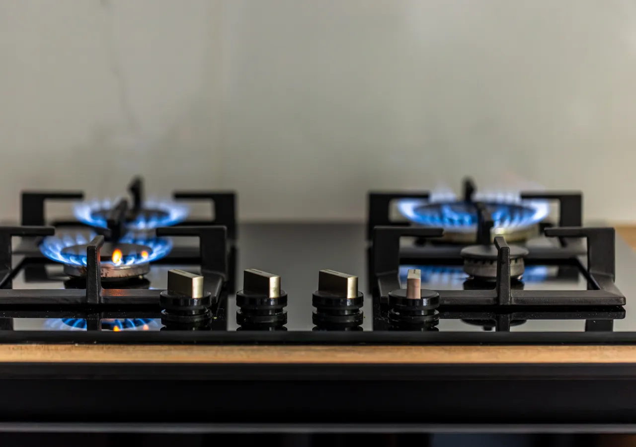 Two burners lit on a modern gas cooktop, the blue flames clearly visible against dark cast-iron trivets and a pale kitchen benchtop behind.