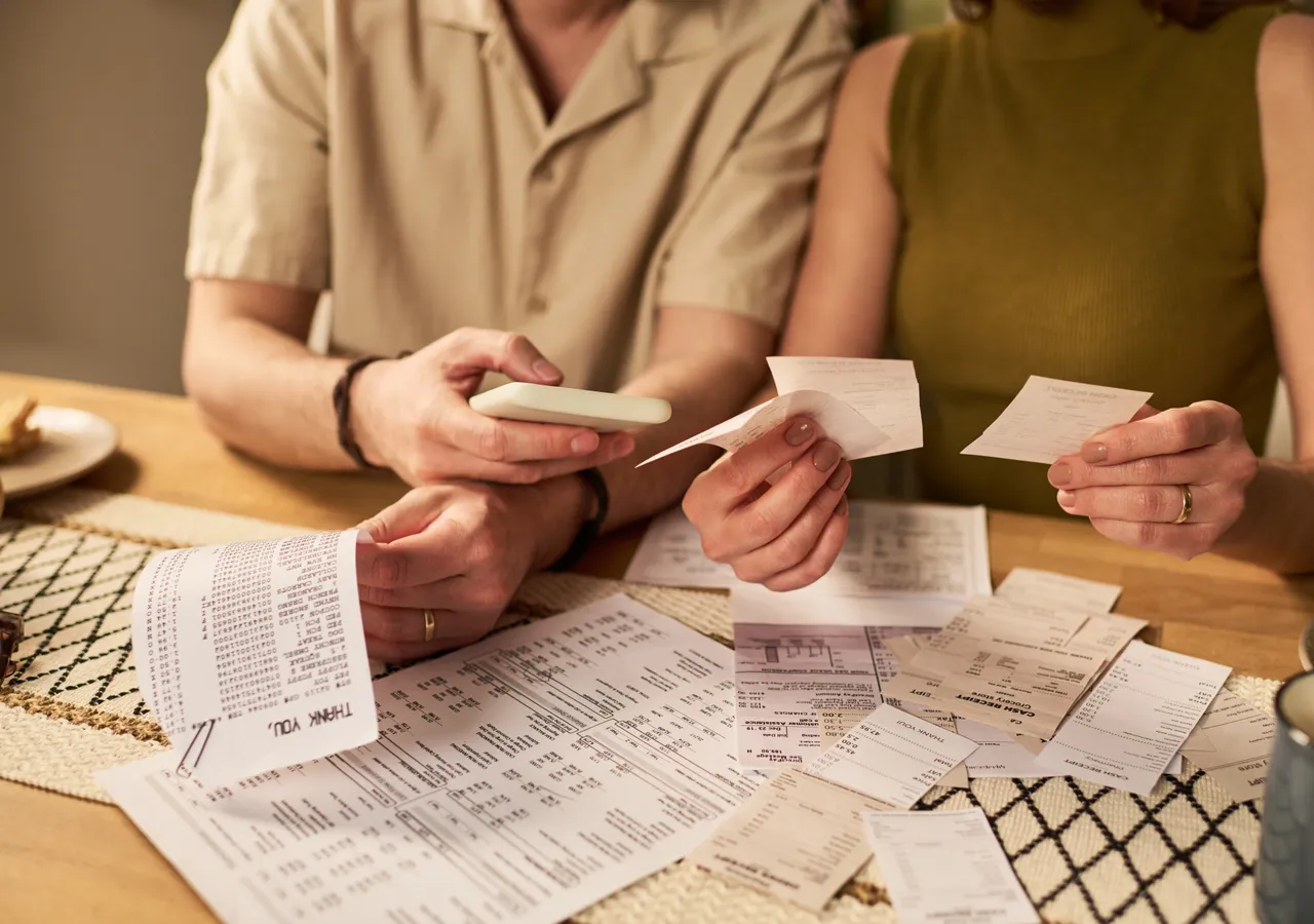 Two people at a kitchen table going through a spread of paper bills and receipts together.