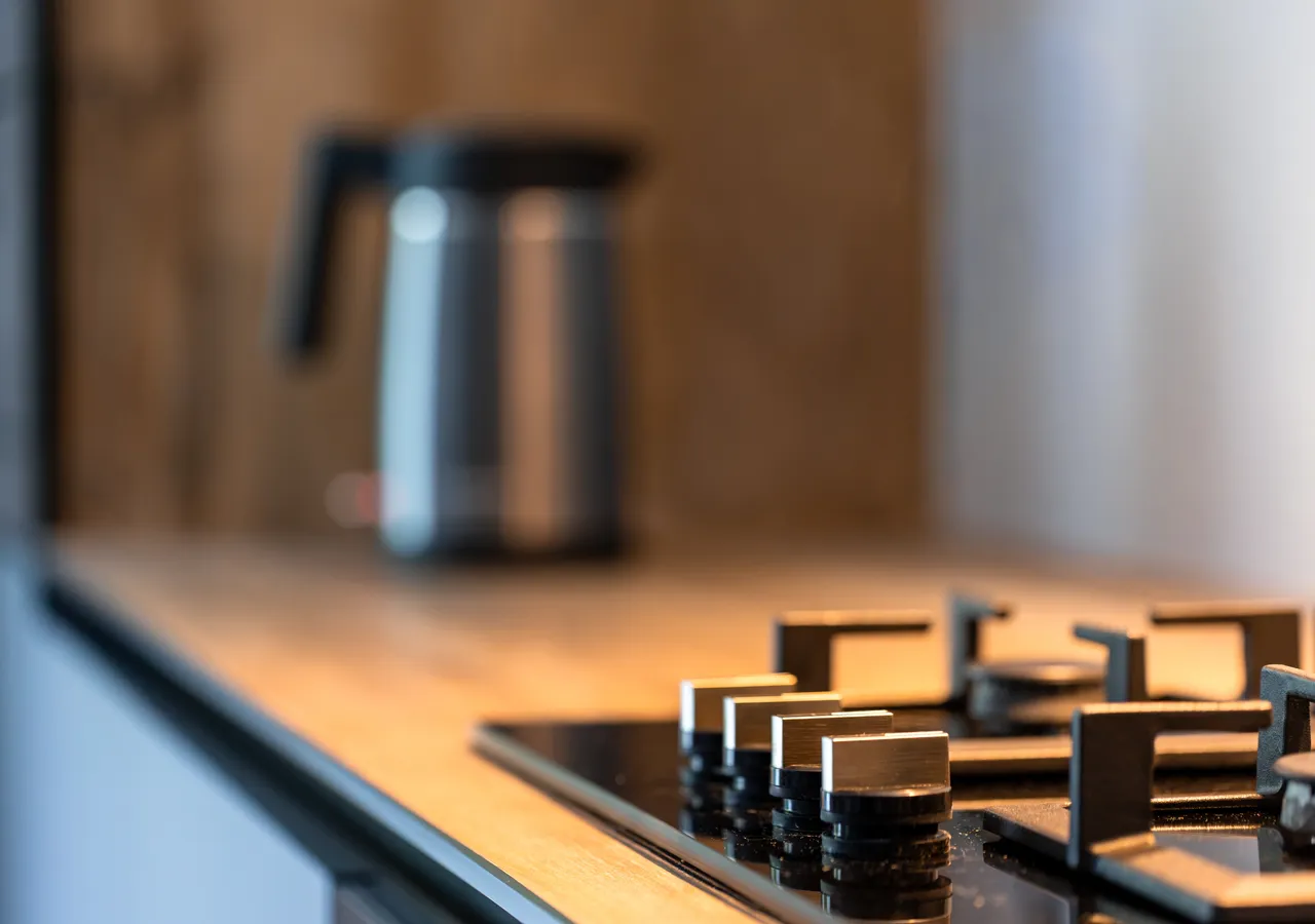 A stainless steel kettle sitting behind the control knobs of a gas cooktop in a dark modern kitchen, shot close and low.