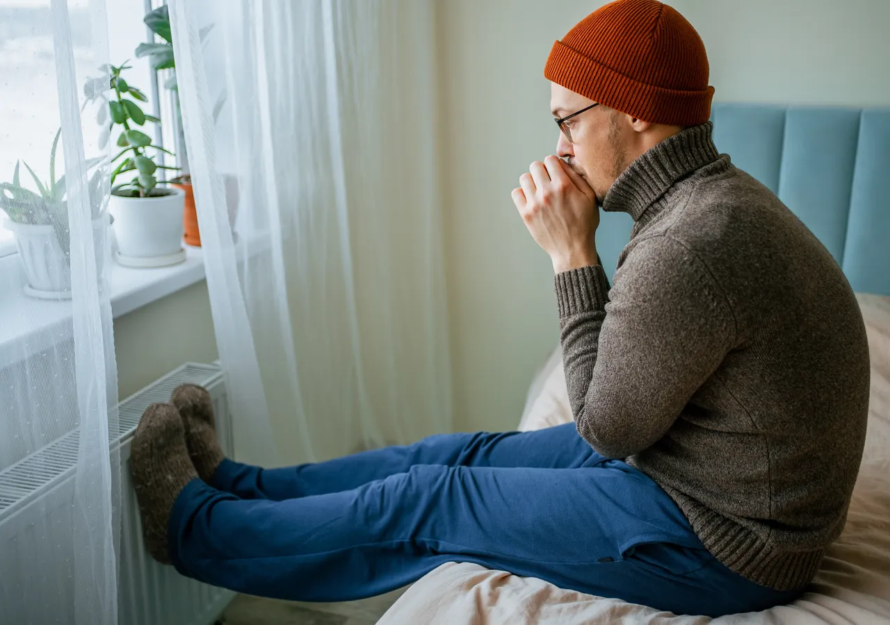 A man sitting on a window seat in a coat and beanie with his hands held to his mouth, in a cold, pale room.