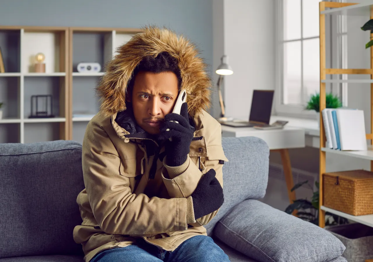 A man in a heavy winter coat and gloves sitting on a sofa indoors, speaking on a mobile phone.