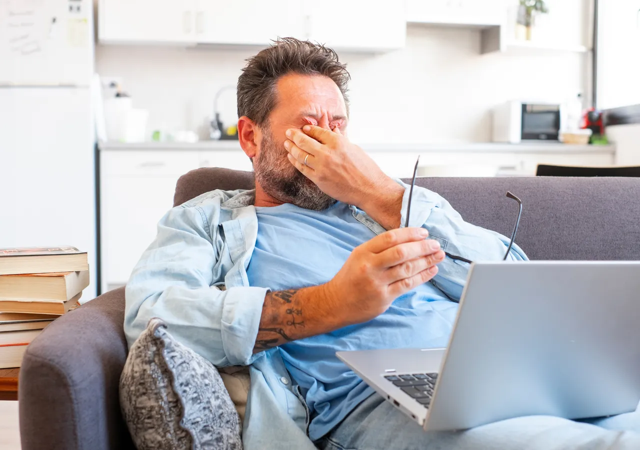 A man sitting on a sofa with a laptop, one hand over his face and a bill in the other hand.