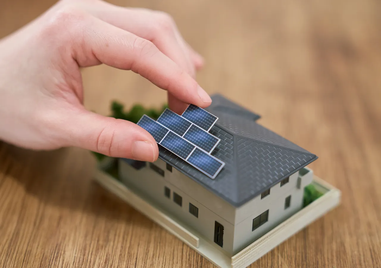 A hand placing a small solar panel onto the roof of a scale model house sitting on a wooden desk.