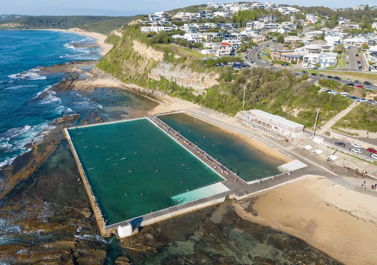 An aerial view of ocean baths beside a headland, with a coastal town rising on the hill behind.