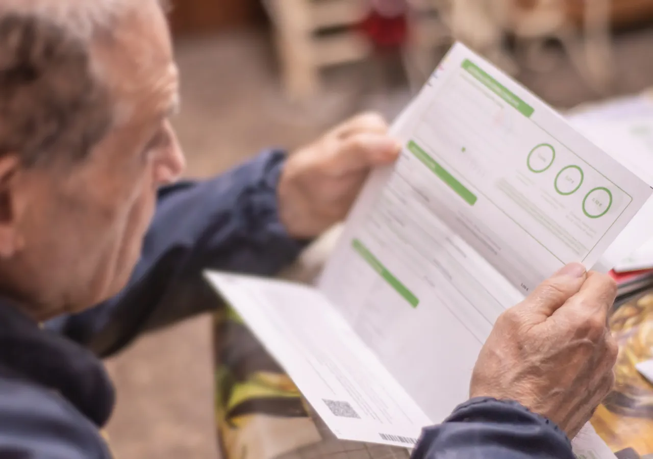 An older man sitting at a table reading through a printed application form, holding a second sheet of paper in his other hand.