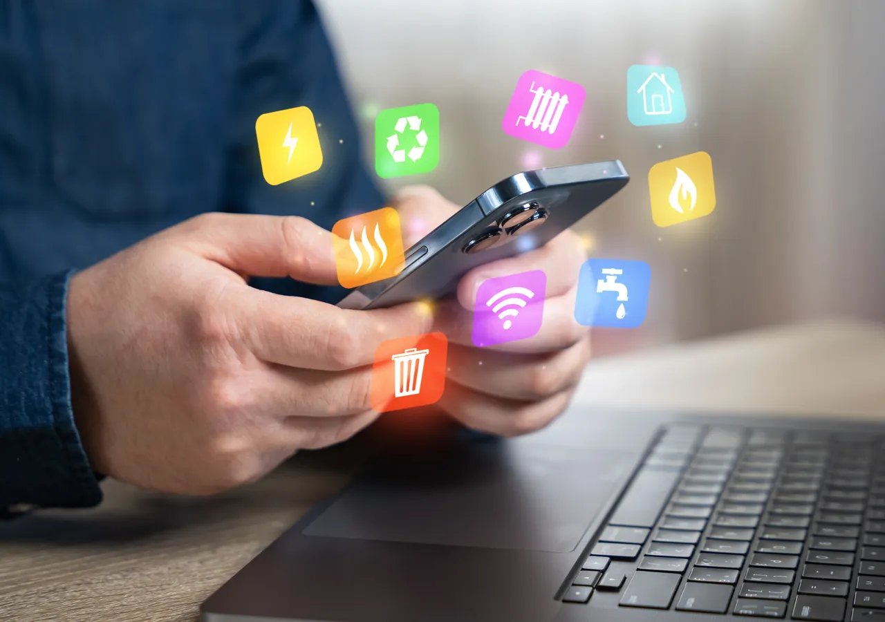 A person holding a phone above a laptop keyboard, with utility and energy icons floating above the screen.