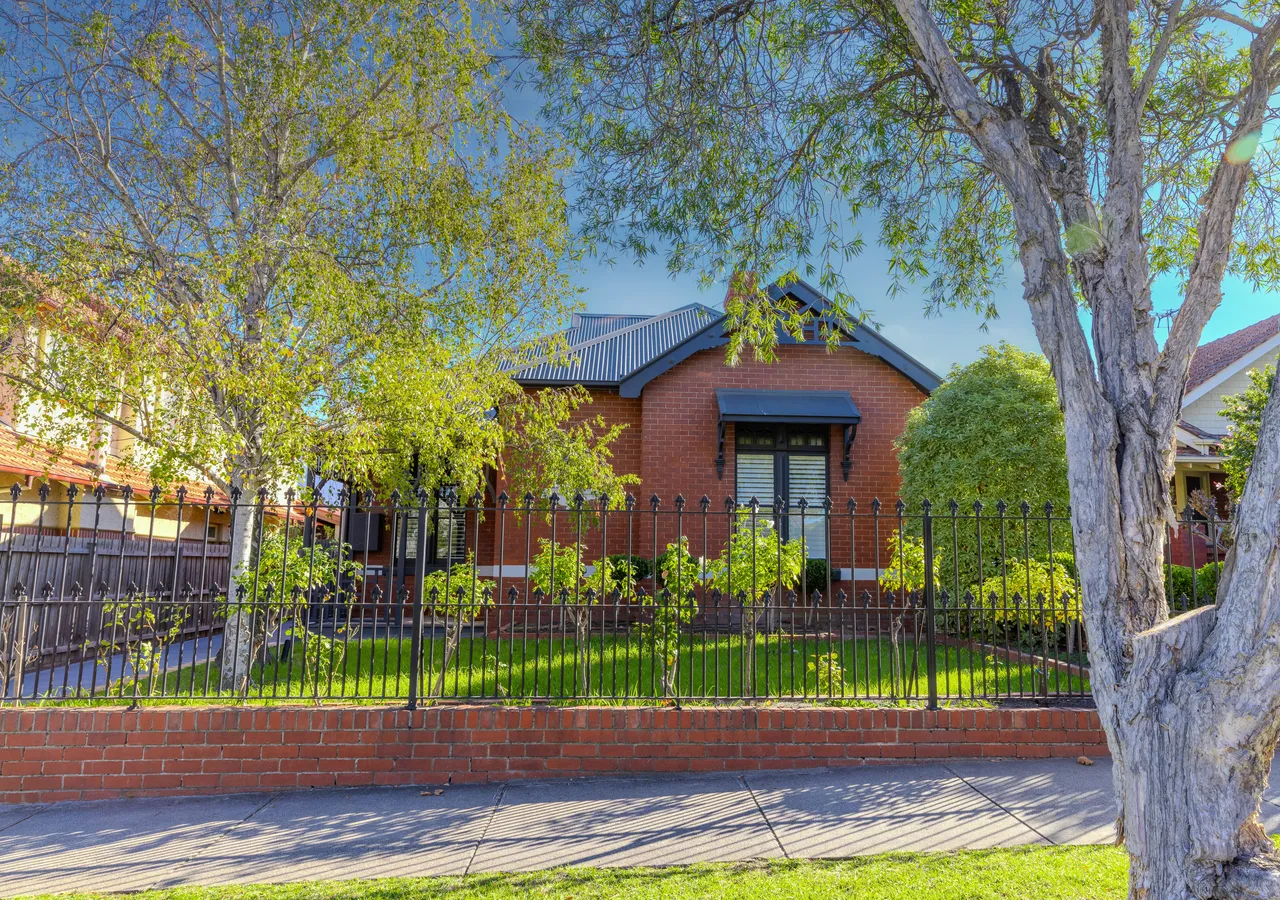 A red brick bungalow with a verandah behind a low fence on a quiet suburban street.