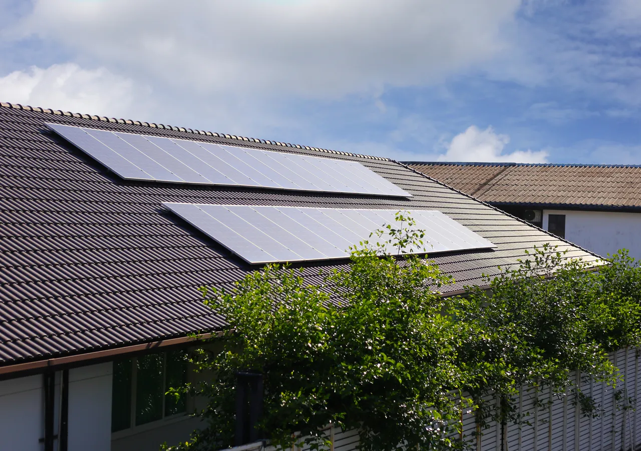 Solar panels installed across the tiled roof of a suburban house, seen from ground level against a blue sky.