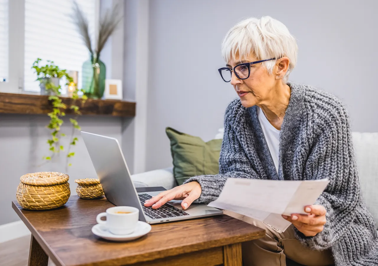 An older woman at a dining table with a laptop open in front of her, holding a paper bill and reading it closely.