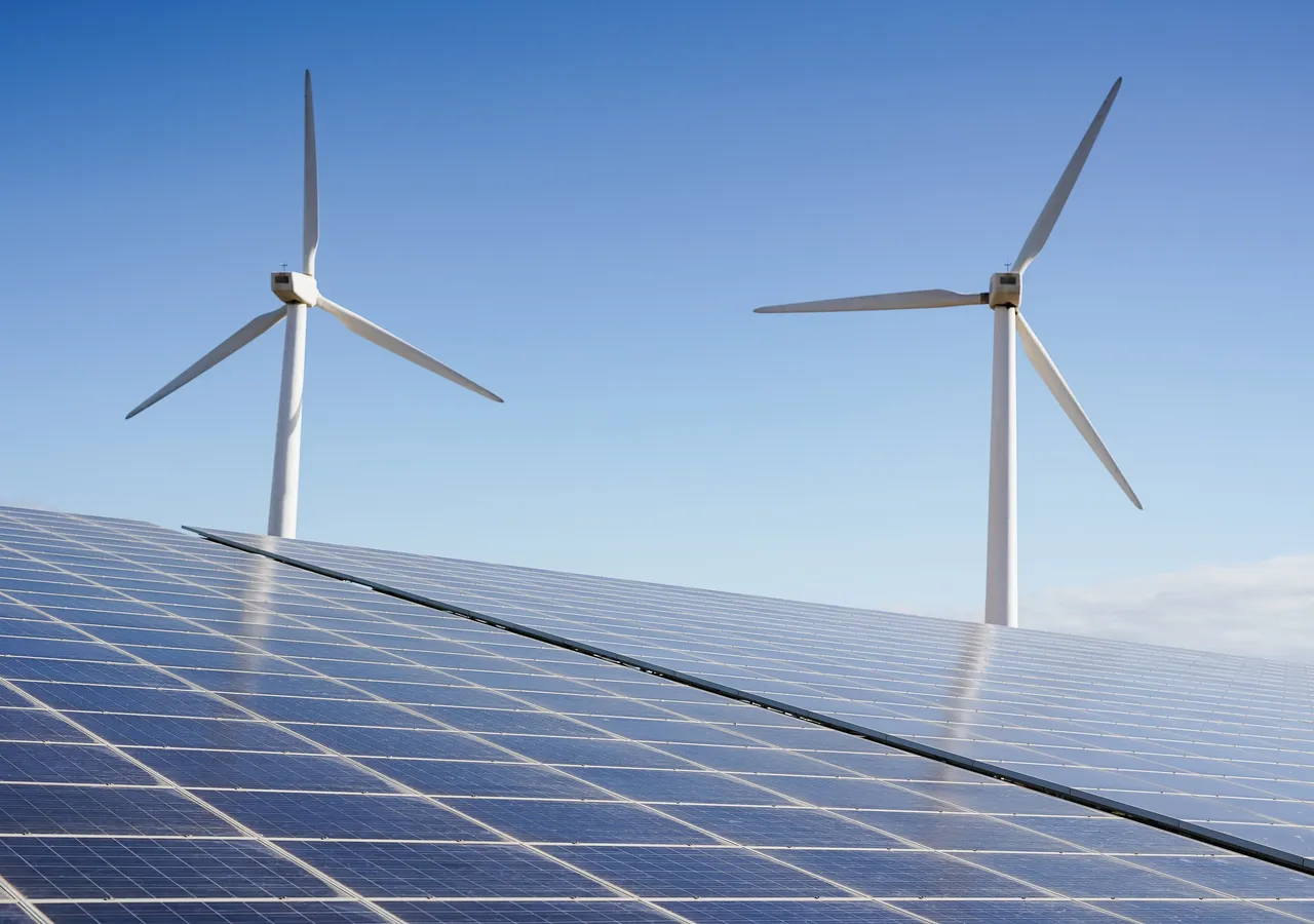 A field of solar panels with wind turbines standing behind them.