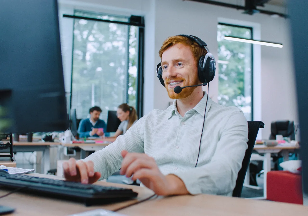 A support agent wearing a headset at a desk in a bright open-plan office.