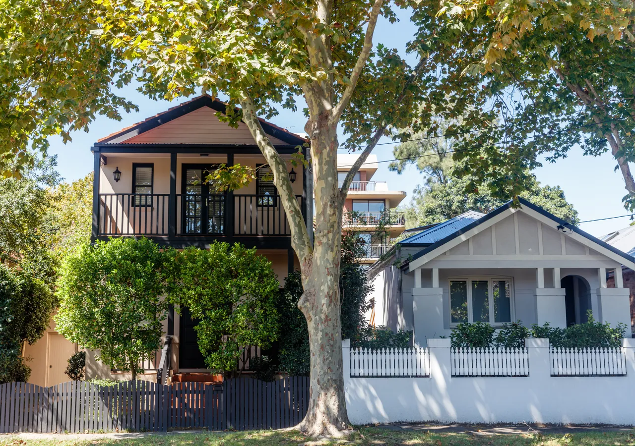 Two inner-suburban Australian homes side by side behind picket fences and a large plane tree: a two-storey house with a balcony, next to a single-storey bungalow.