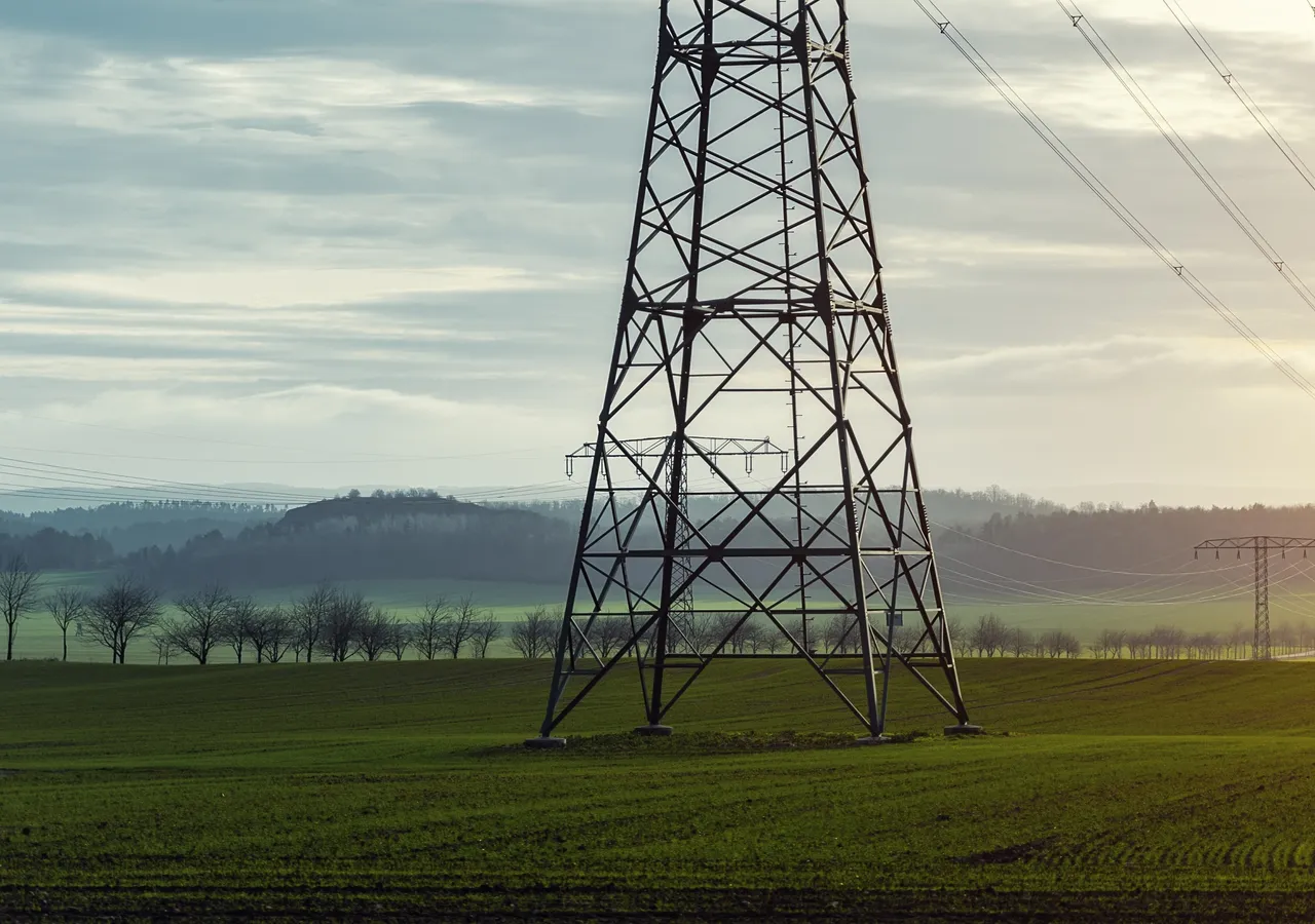 A high-voltage transmission tower carrying power lines across open farmland at sunrise.