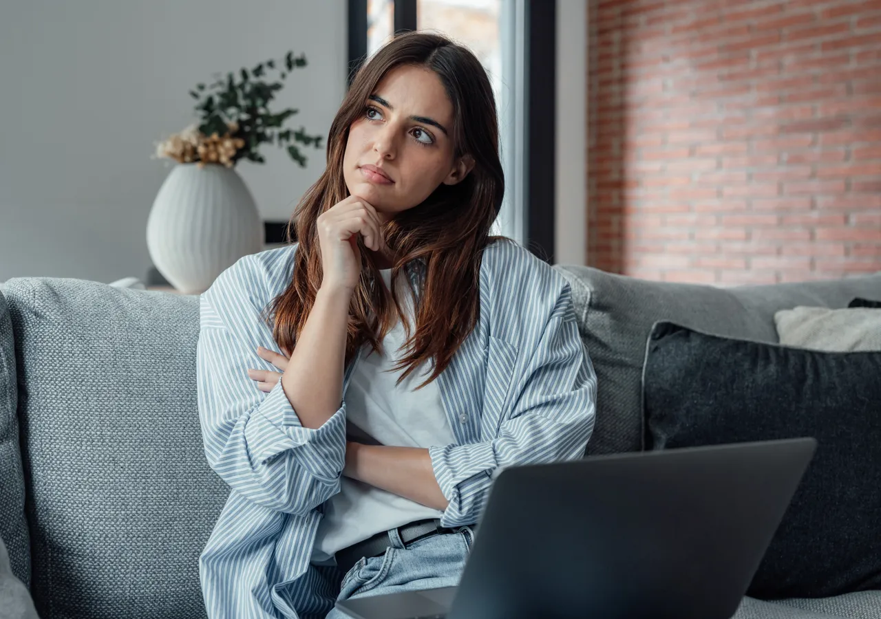 A woman sitting on a sofa with a laptop on her knees, resting her chin on her hand as she reads the screen.