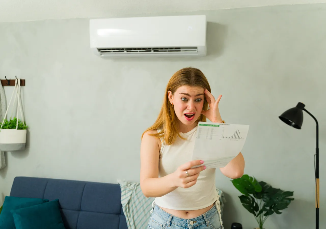 A woman standing in a living room reading a bill with a surprised expression, a wall-mounted air conditioner visible above her.